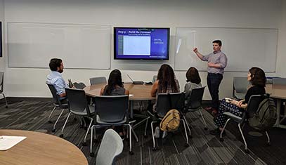 man standing and gesturing towards presentation screen with small group